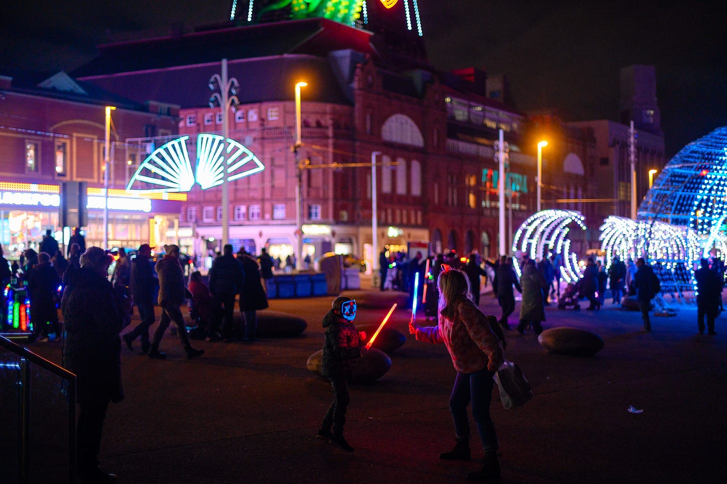 A kid engages in a lightsaber battle with a grownup at night during Blackpool Illuminations.
