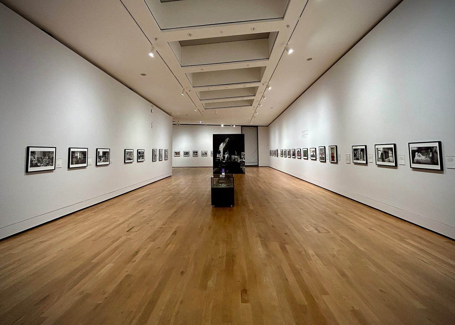 This photo showcases the 'Where Ideas Are Born' photography exhibition at Aberdeen Art Gallery. The gallery space is wide and welcoming with a polished wooden floor leading to a central dark panel, creating a corridor-like effect. Both sides of the walls are lined with framed black and white photographs, evenly spaced and illuminated by ceiling lights. The exhibit seems to offer a quiet, contemplative atmosphere, inviting visitors to immerse themselves in the visual storytelling of the photographs.