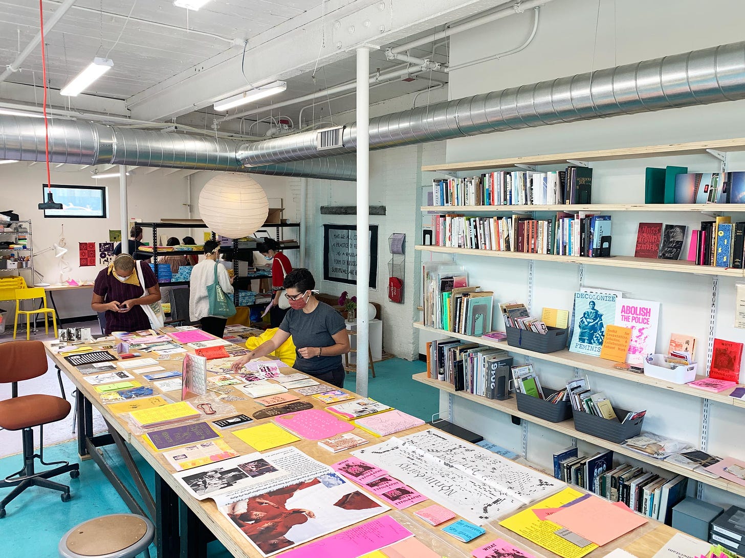 View of the Queer.Archive.Work library and reading space, showing a colorful array of printed materials laid out on long tables. On the wall are shelves of books and zines. Several masked people are in the space looking at the materials.
