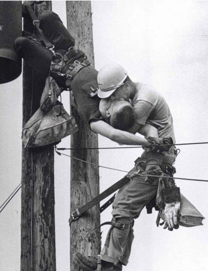 A lineman up on a pole giving mouth-to-mouth resuscitation to another lineman who appears to have been electrocuted and is dangling lifelessly in the air..