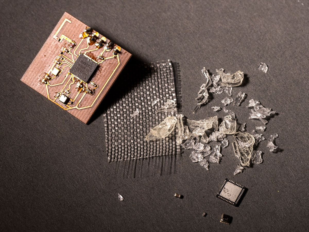 A small brown circuit board sits on a gray background. To its right are a small copper plate, sheets of glass fibers in a crosshatch pattern, small chunks of vitrimer plastic that’s been removed from a circuit board, and a computer chip