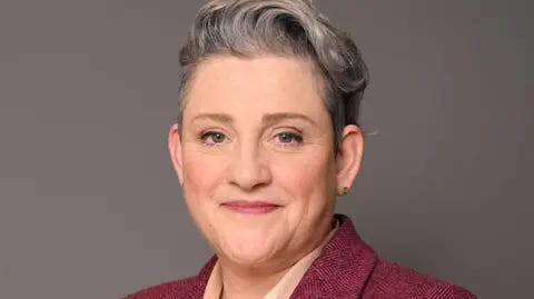 UK Parliament Head shot of Ashley Dalton in front of a plain, dark grey background. The MP is smiling and has short grey hair which is longer on top. She is wearing a burgundy suit jacket and a cream blouse.