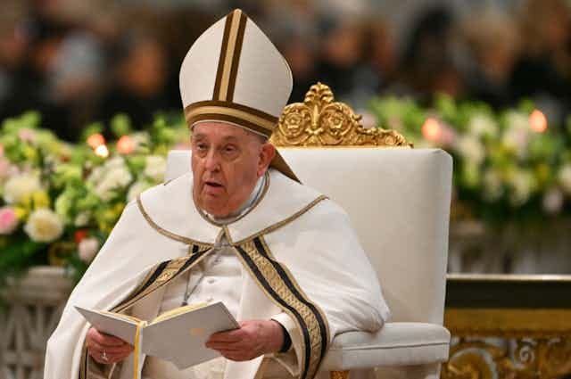 A man in white mitred hat and cloak, seated on a white and gold chair, holding a document or book