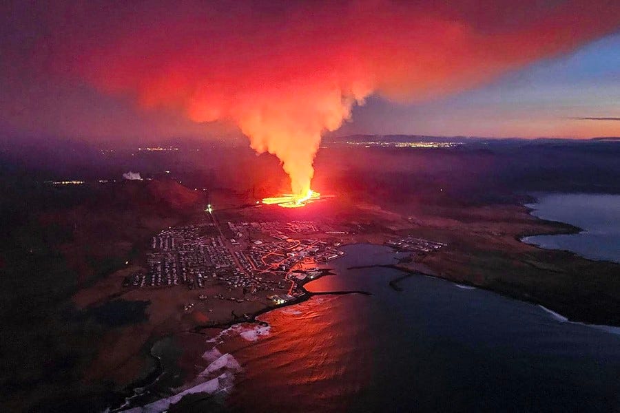 An aerial view of a coastal Icelandic town with an erupting volcanic fissure a short distance inland, just uphill from its houses