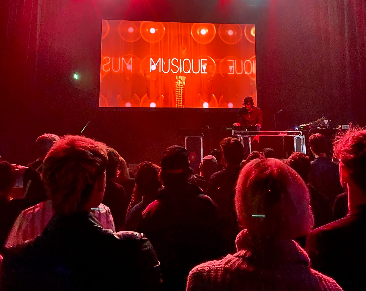 Palco com um rapaz manipulando uma mesa de som, um telão atrás vermelho escrito MUSIQUE e a plateia à frente.