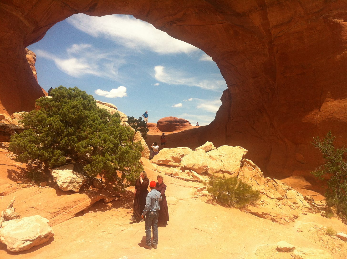 Two FLDS women, in long black dresses, talking to a man in jeans. A large red rock formation with an opening to the sky is in the background.