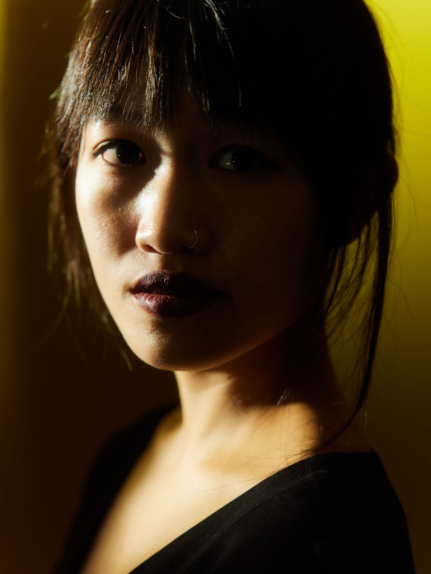 A portrait of a woman with dark hair and bangs, wearing a nose ring and dark lipstick. The lighting casts shadows on half of her face, creating a dramatic contrast against a soft yellow background.