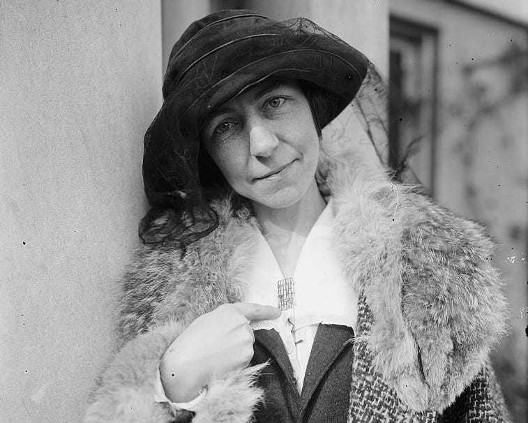 Black-and-white portrait photograph of Julia Emory, a white woman who wears a fur coat and veiled hat. She is pointing to a pin on her lapel in the shape of a prison cell gate.