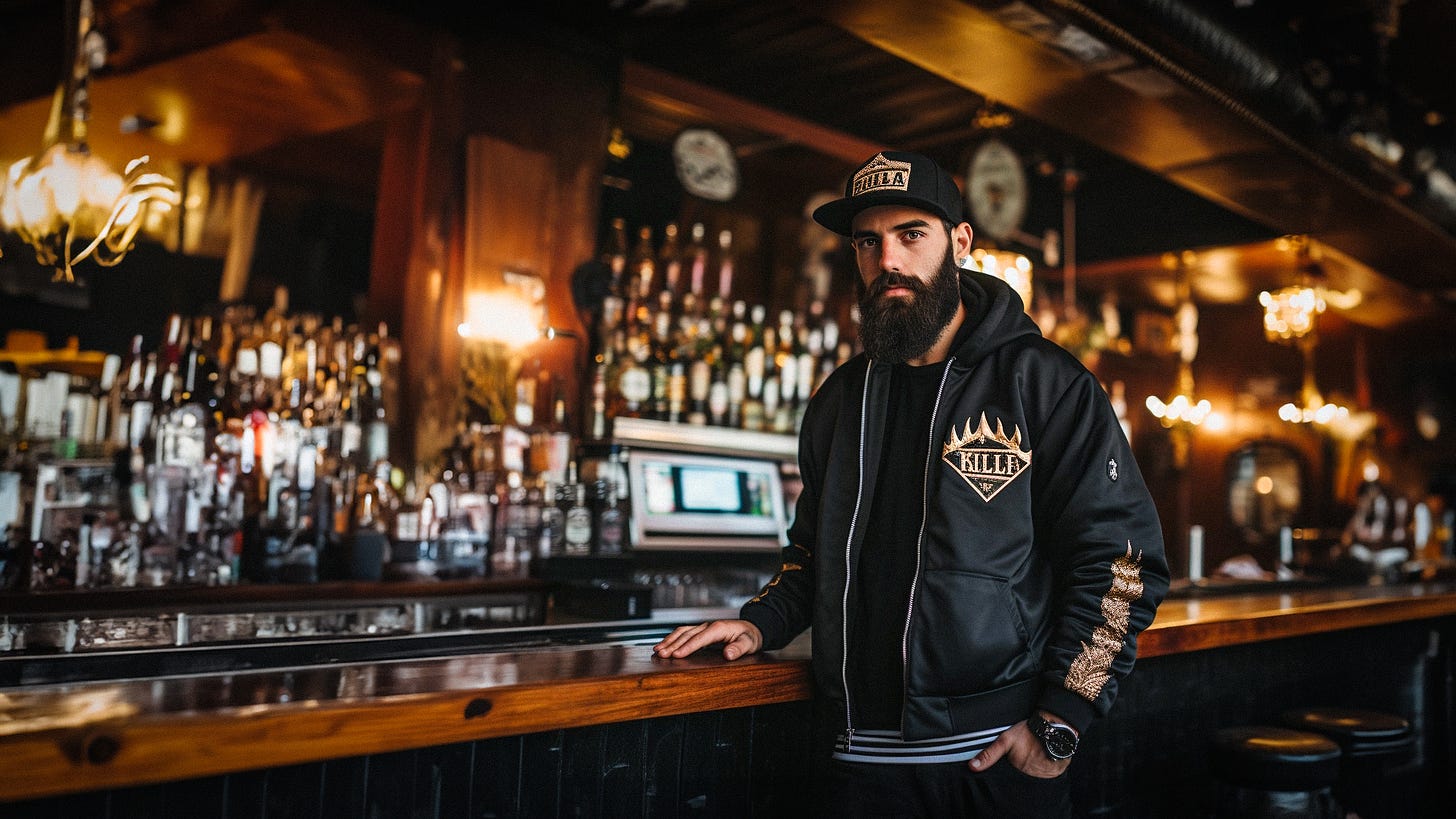 A man with a long beard stands at a bar with a large display of alcohol behind him next to a point of sale. The man wears an embroidered bomber jacket and an embroidered flatbill cap. 