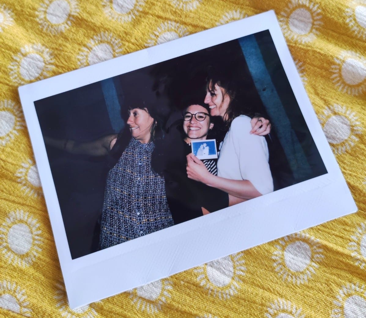 Polaroid con tre amiche su uno sfondo giallo. Da sinistra a destra: una persona girata di lato che sta chiamando altre persone, una persona che guarda in camera abbracciata a una terza persona che sta ridendo.