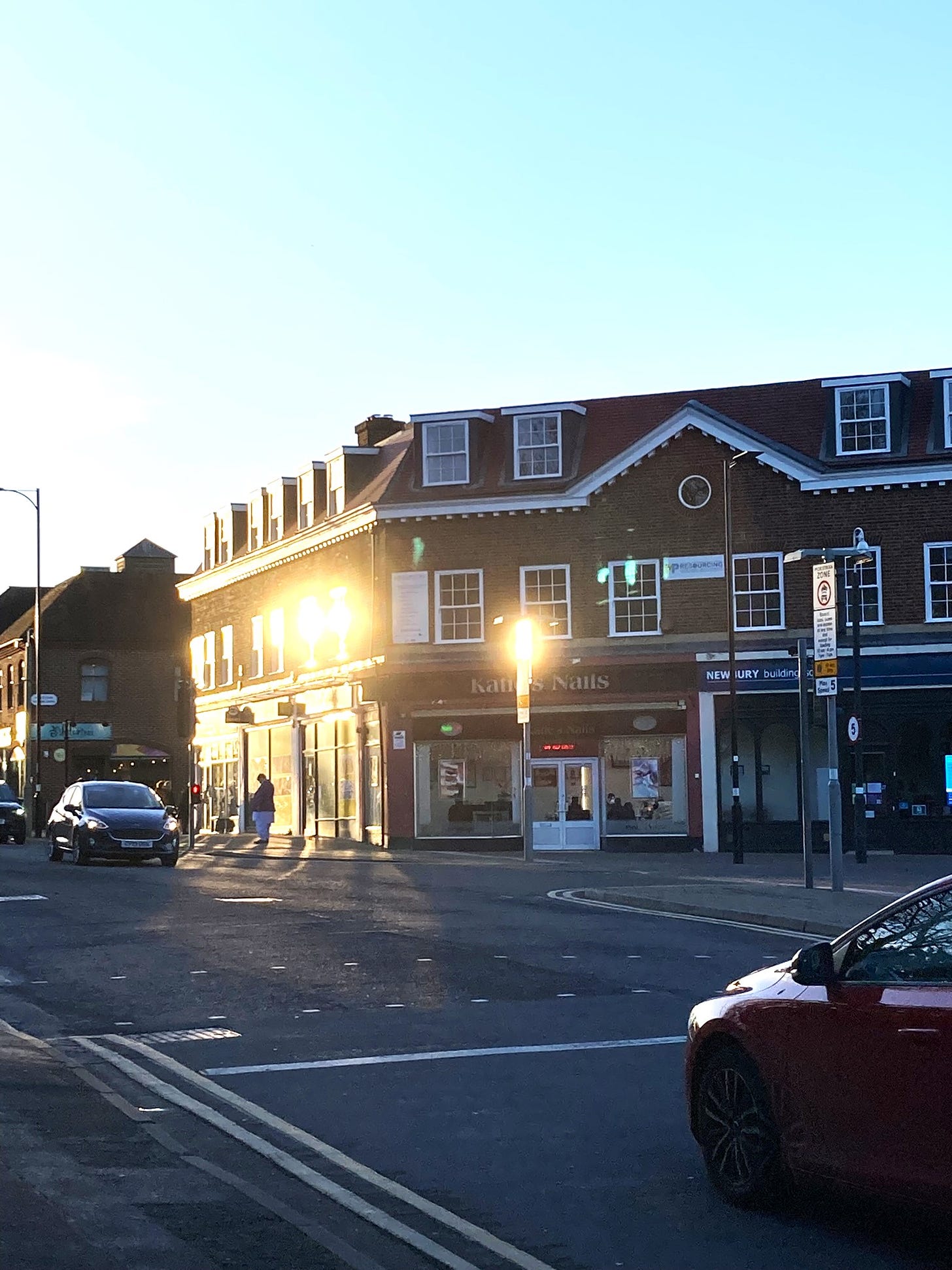 An average high street featuring shops, nail salons, building societies and cars. The sun is glinting off the windows, under a bright blue sky.