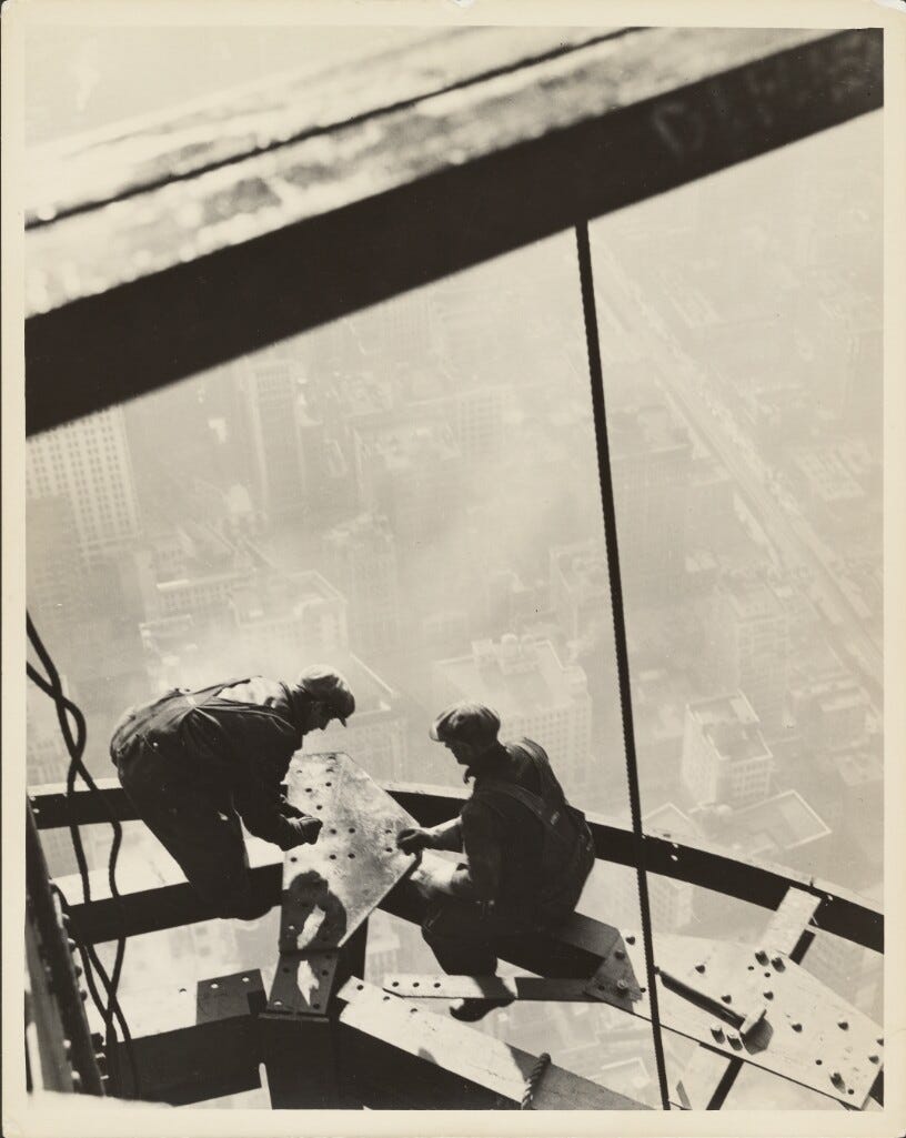 Black and white photo of two men bent over putting rivets into part of the Empire State Building far above the ground