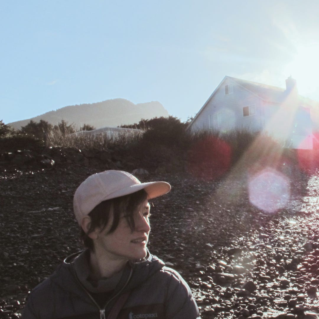 Sarah looks off to the side, standing on a rocky shore in front of a wooden house. The sun is at their back. She wears a jacket and a pink baseball cap.