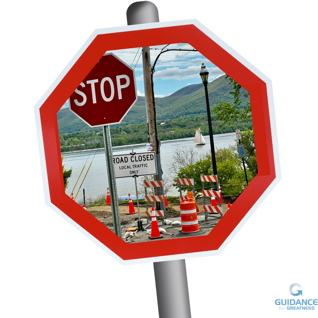 A scene of a road closure, complete with signage and orange cones with the Hudson River and Mount Beacon behind. A beautiful sloop sails the Hudson. The photo is framed as an octagon and is superimposed on a red stop sign that is leaning slightly to the left.