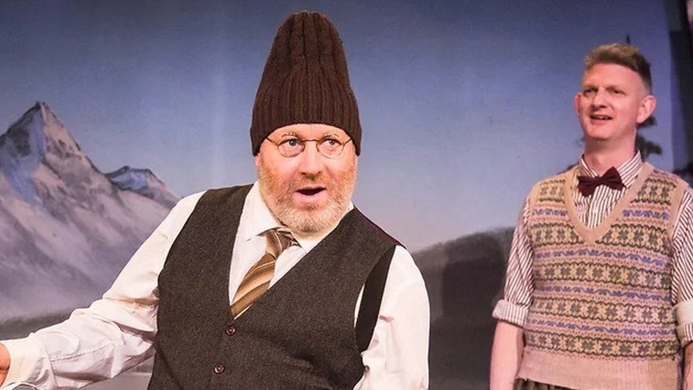 Howard Coggins on stage wearing round glasses, with a beard, white shirt and a brown tie, waistcoat and a woolly hat on. Stu Mcloughlin is behind him