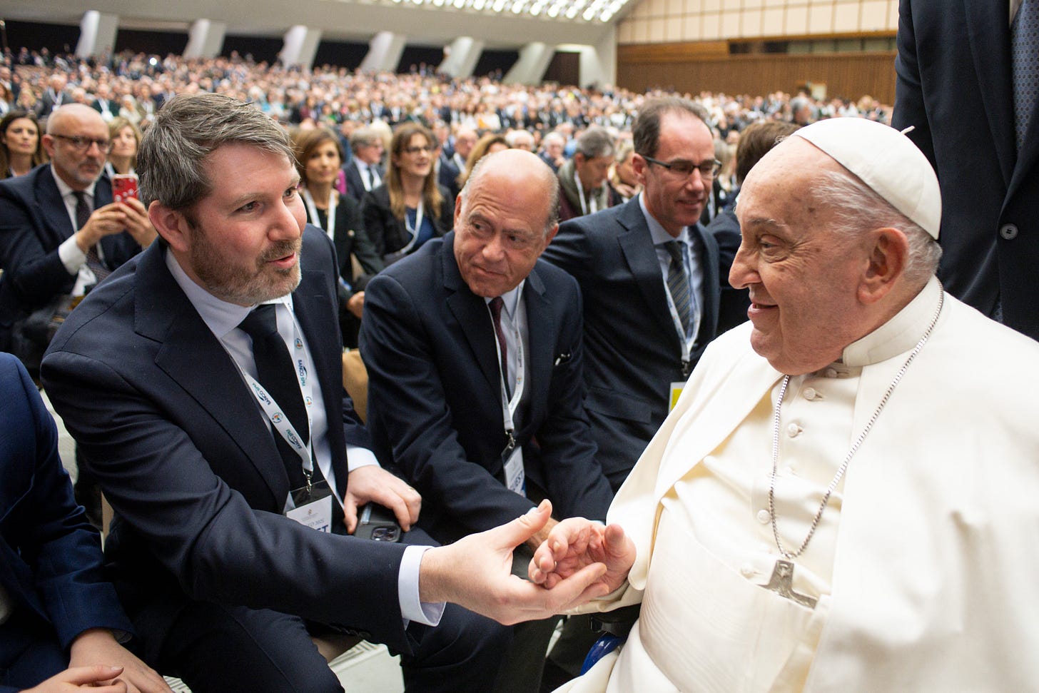 At left, a seated, suited middle-aged man with salt and pepper hair grasps the hand of the seated man at right, Pope Francis