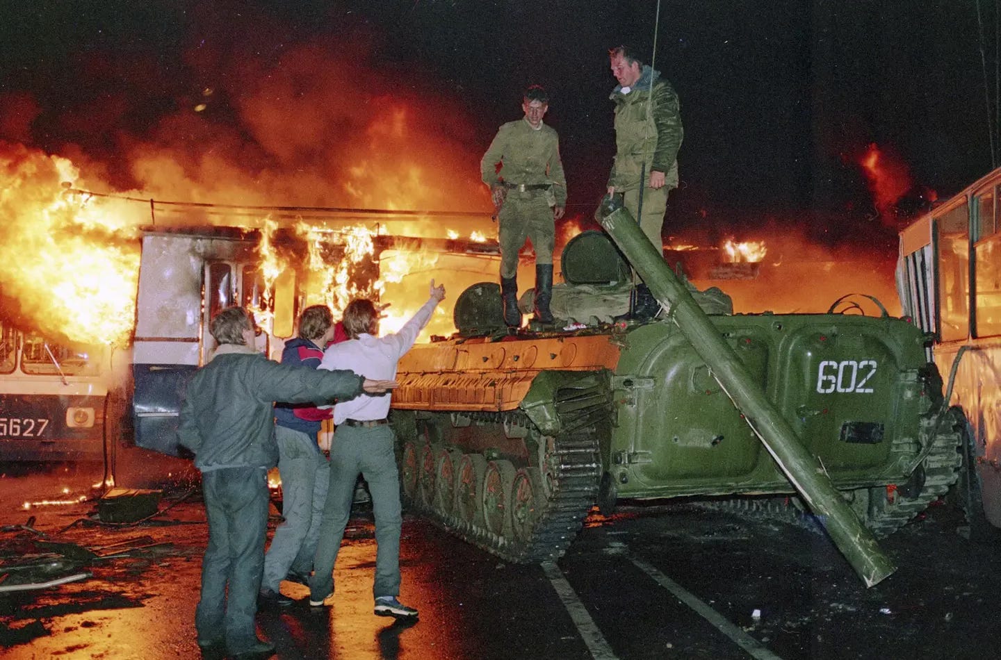 A tank that has crashed into a burning bus. Soldiers are on the tank and protesters are shouting at the soldiers.