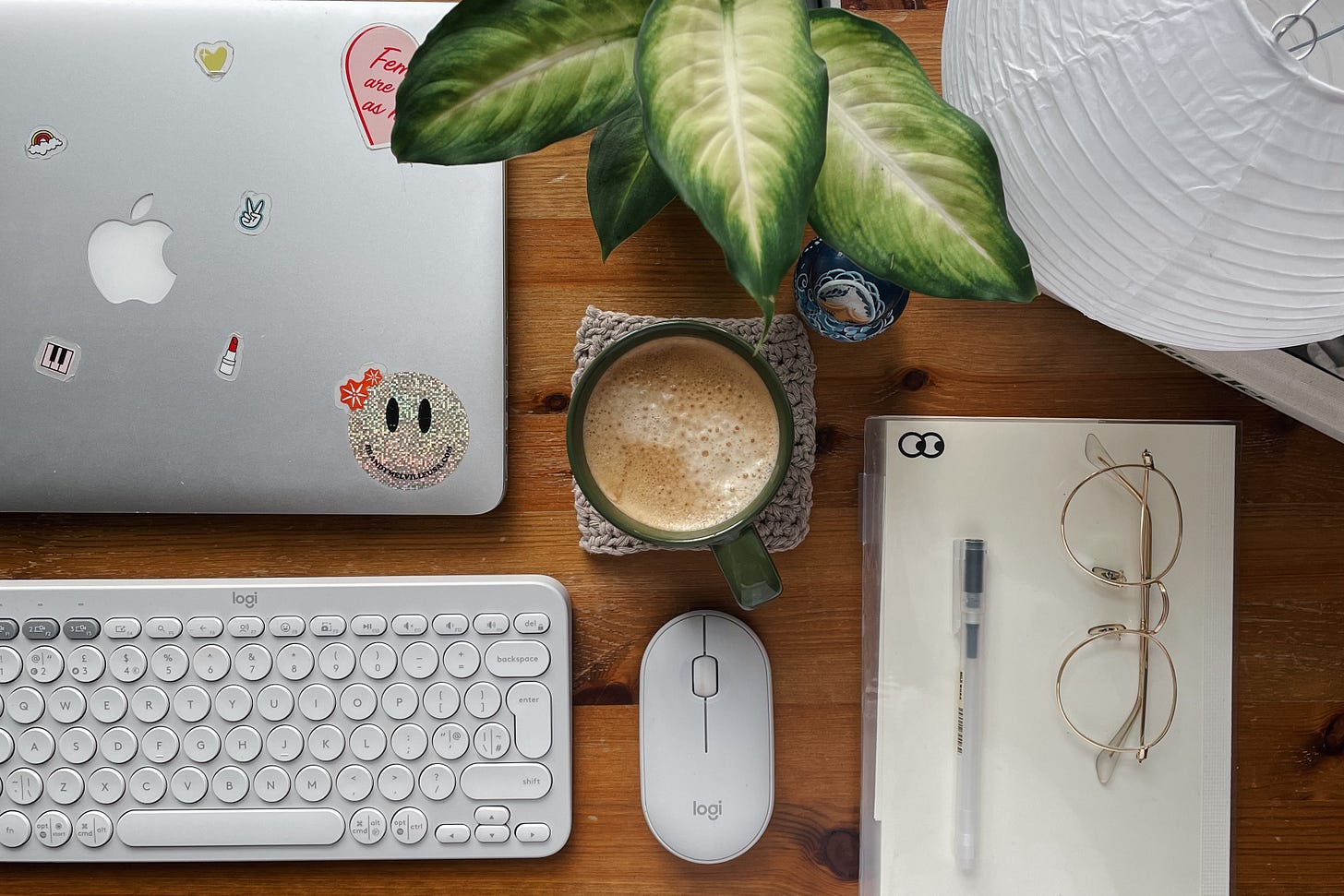 Flatlay of a wooden desk with a laptop, keyboard, mouse, notebook, glasses, and a freshly brewed coffee. A green plant adds a touch of life