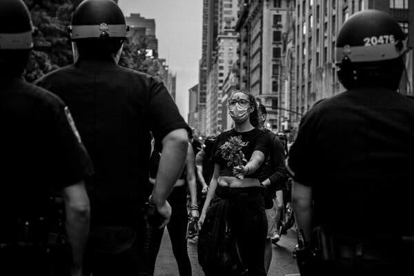 On a New York City street, with buildings in the background, a masked protester in a T-shirt holds out her left arm toward police, whose backs are toward the viewer. 