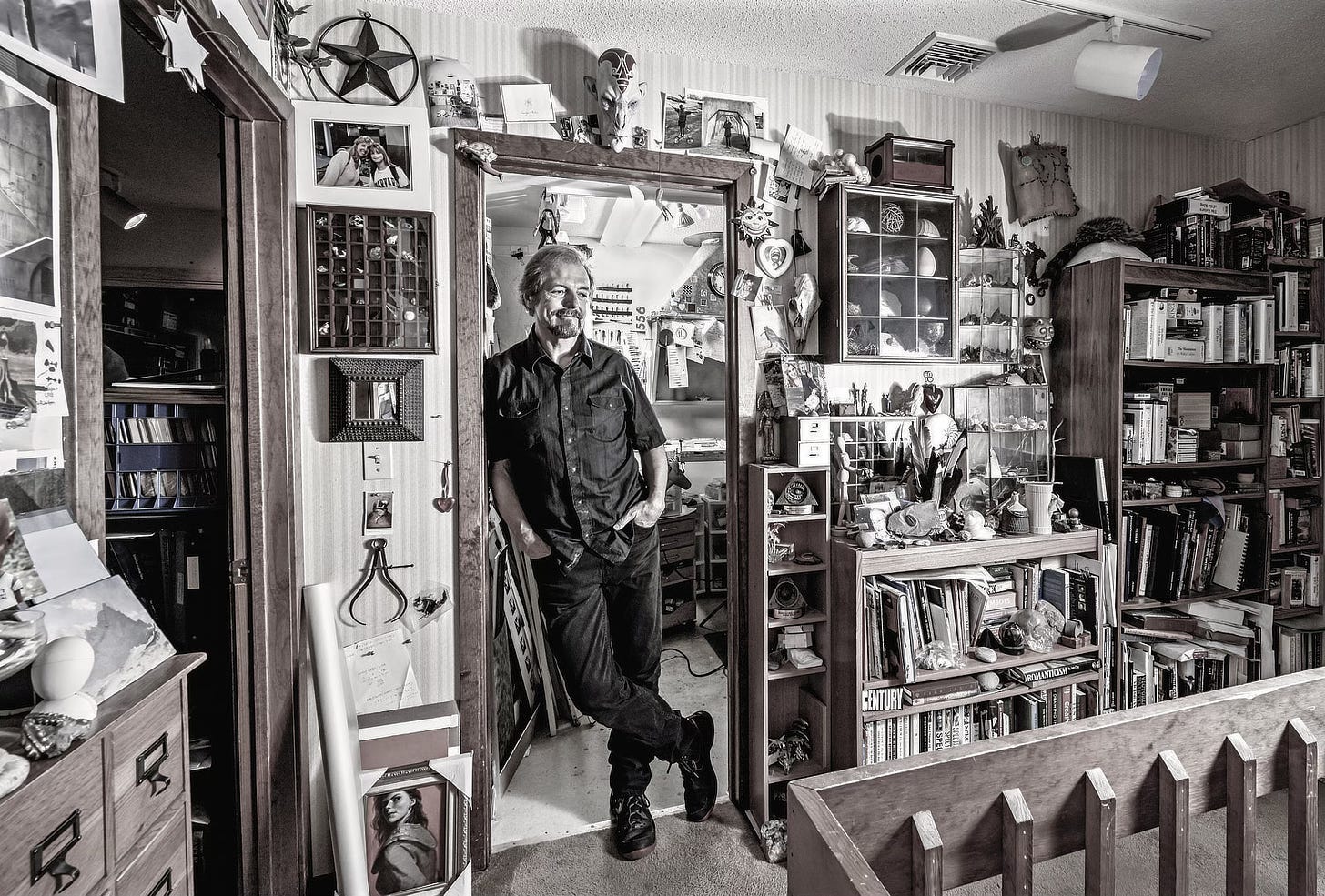 Sepia tone high definition photograph of Michael Whelan leaning against the doorway in his studio. An assortment of knick knacks decorate the walls. To the right, shelves are full of art books.