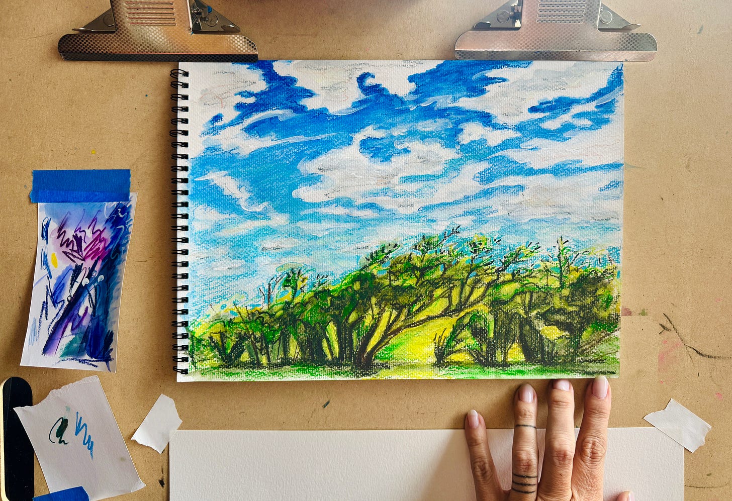 Watercolor and artist crayon illustration made en plein air featuring bright blue skies, wavy clouds, hot yellow and green foliage on a wire bound watercolor notebook clamped to an artboard.