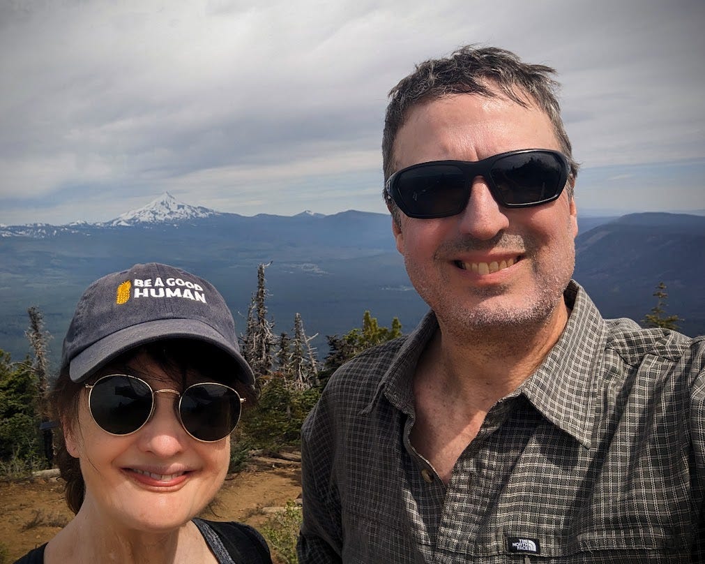 Man and woman standing on top of a mountain smiling with gray clouds in background and pine trees behind them.