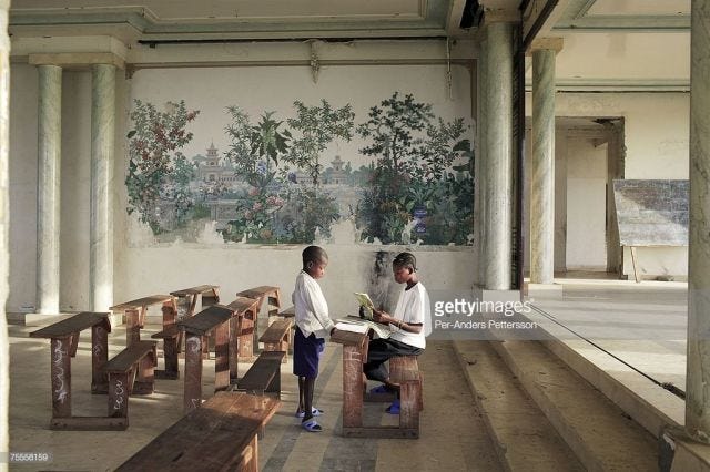 Two young boys sit at wooden desks inside an ornate, palatial room