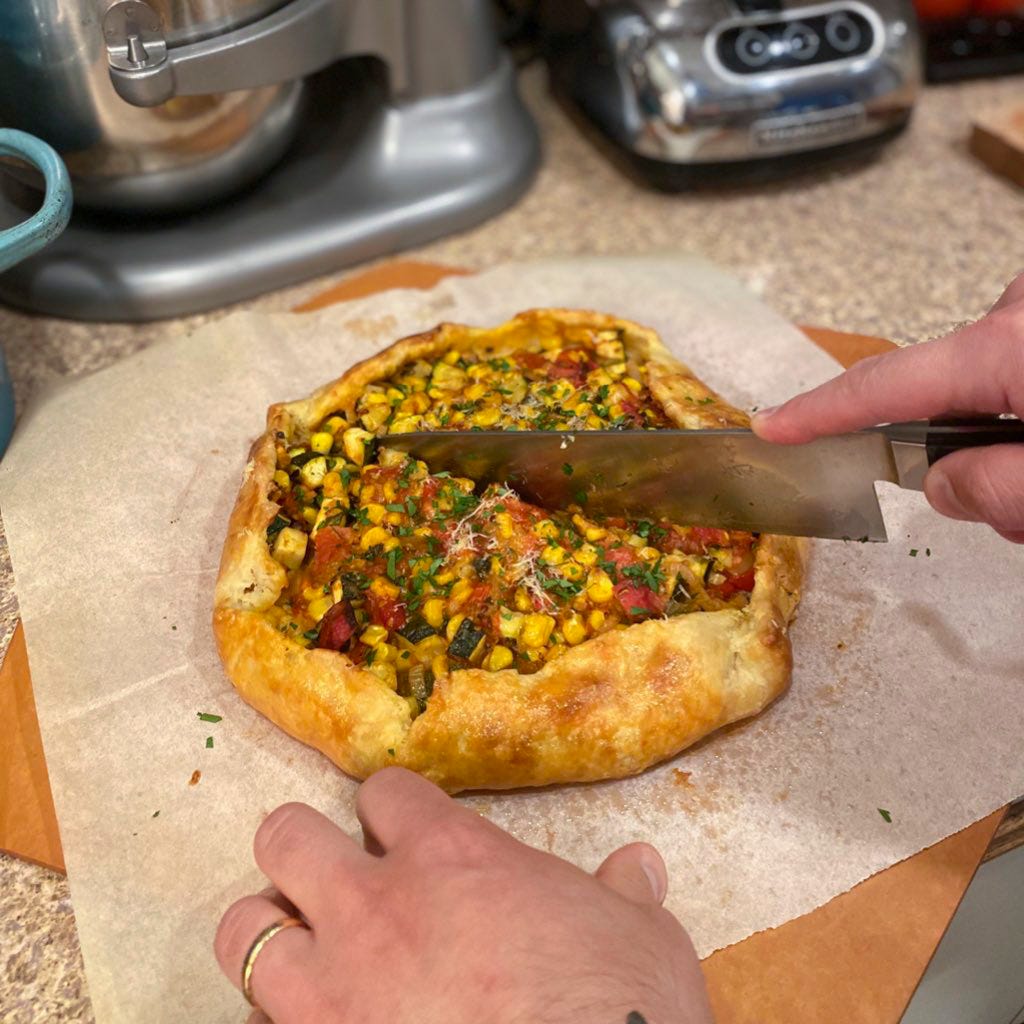 Jeff is cutting the galette described above with a large chef's knife. His other hand holds the parchment it's resting on still. The galette is browned at the edges, and the centre is filled with corn, tomato, and zucchini, with grated parmesan and chopped parsley on top.