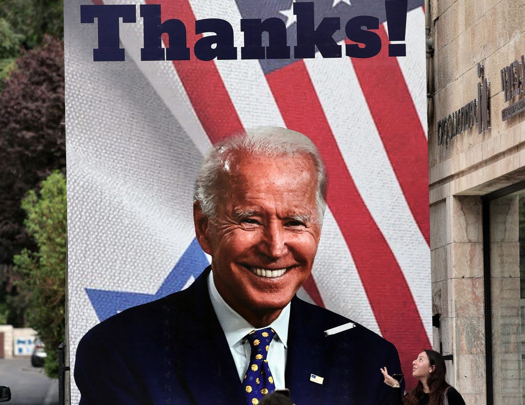 A giant poster of Biden, grinning in a dark suit, smile lines surrounding his eyes, fills a street. The poster's background features the US and Israeli flags,. A dark haired woman in black clothes blows a kiss at the poster.