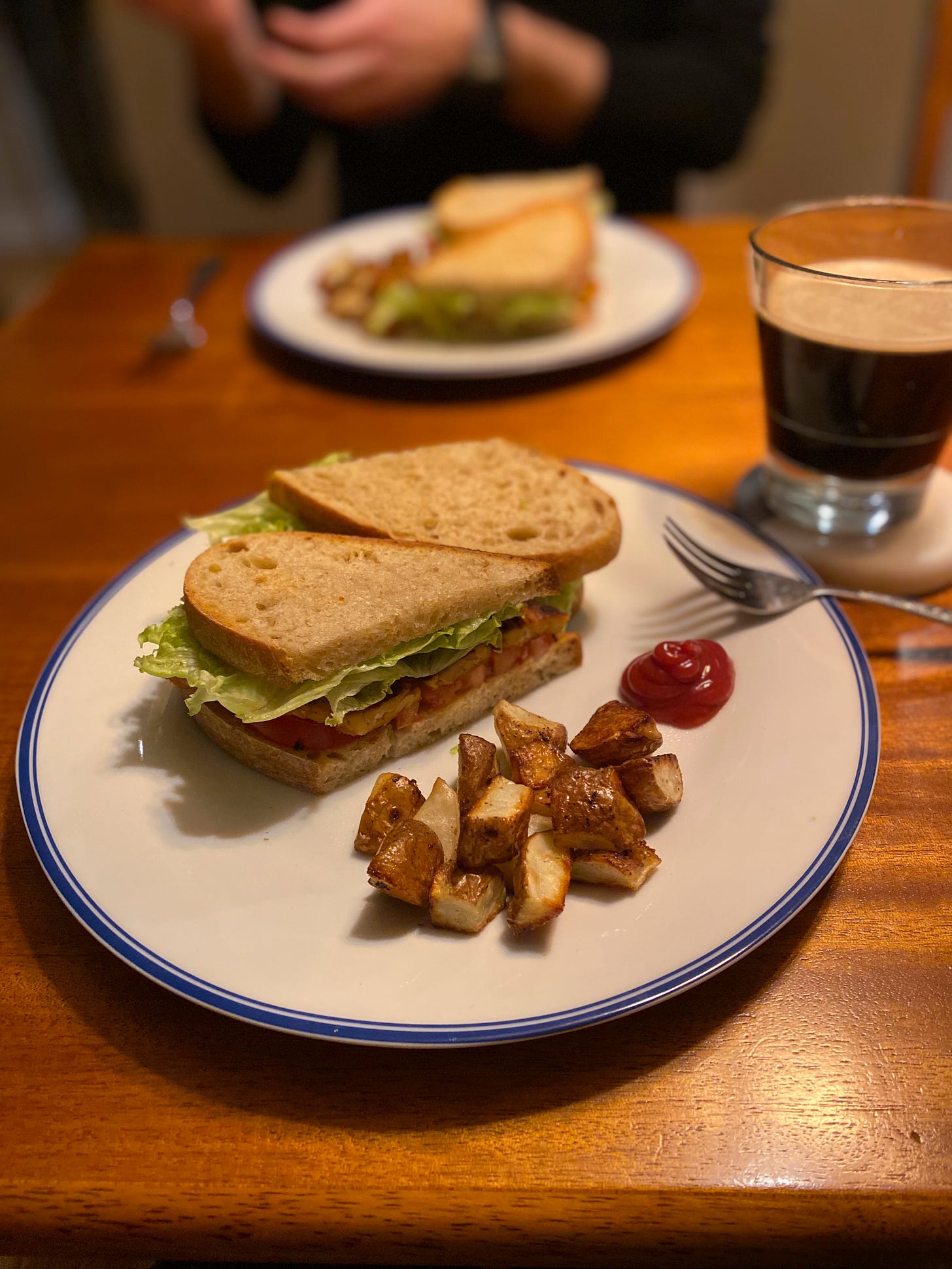 A large white plate with a blue rim holds a small pile of oven fries next to a blob of ketchup, and two halves of a tempeh BLT. Next to the plate on a coaster is a glass of dark beer. Jeff's hands are above his plate in the background, out of focus.