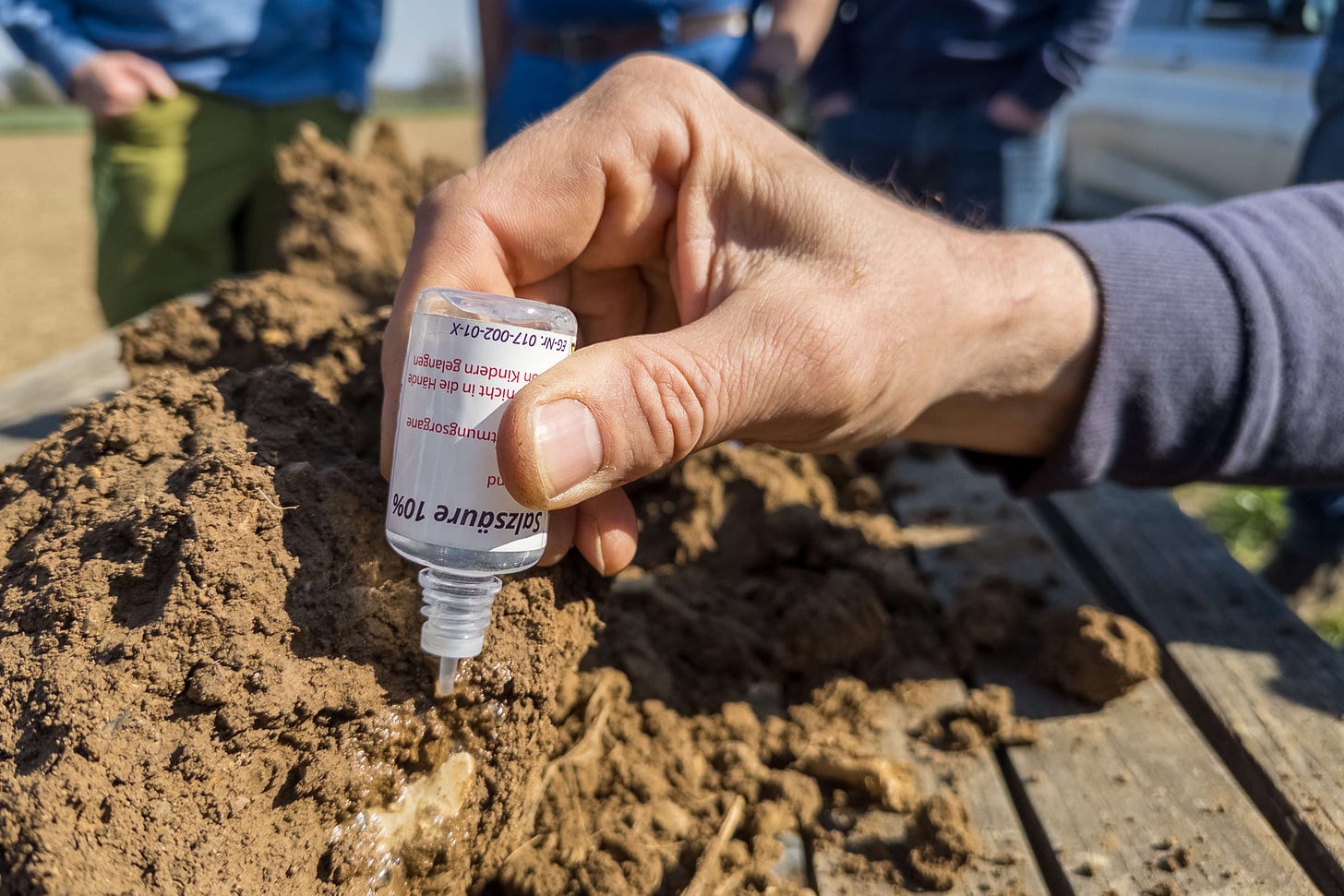 Eine Hand träufelt verdünnte Salzsäure auf eine Bodenprobe, um den Kalkgehalt zu bestimmen – eine einfache Methode zur Analyse der Bodenbeschaffenheit in der regenerativen Landwirtschaft. Im Hintergrund beobachten weitere Personen den Vorgang. Eine Hand träufelt verdünnte Salzsäure auf eine Bodenprobe, um den Kalkgehalt zu bestimmen – eine einfache Methode zur Analyse der Bodenbeschaffenheit in der regenerativen Landwirtschaft. Im Hintergrund beobachten weitere Personen den Vorgang.