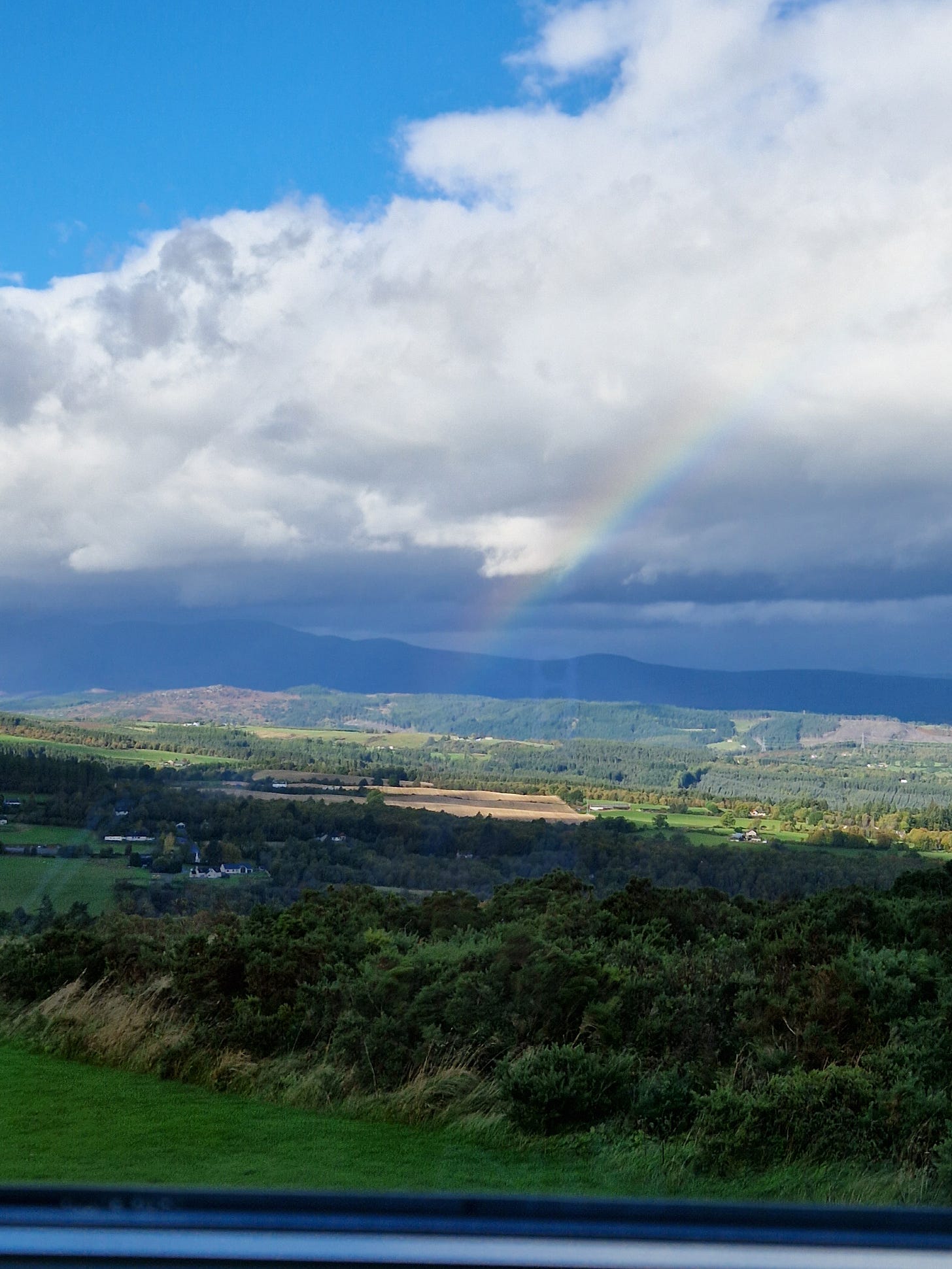 The view over Kiltarlity, with a blue sky, white and grey clouds, and a brief view of a rainbow. 