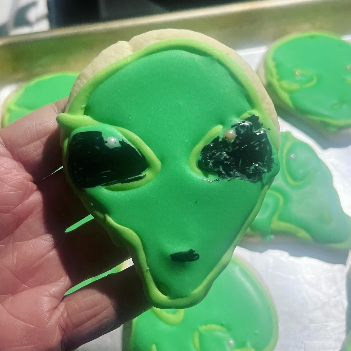 An alien-head shaped sugar cookie topped with royal icing in two shades of green. The alien has black eyes with a round sprinkle as a highlight.
