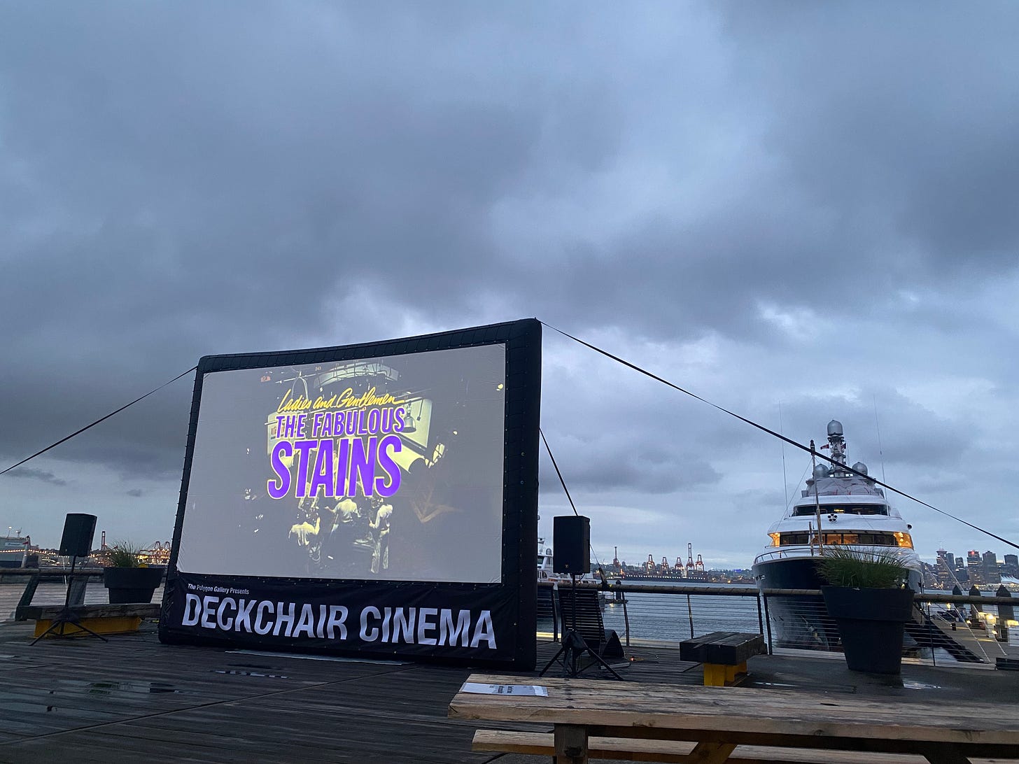 an area of the Lonsdale pier where a screen has been set up with the banner 'DECKCHAIR CINEMA' beneath it. The screen shows the titlecard for 'Ladies and Gentlemen, The Fabulous Stains'. In the background a large boat is docked, with a glimpse of the city in the background, beneath a cloudy sky.