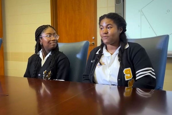 Photo of two young Black high school students wearing matching school uniform sweaters. They are seated at a table, there is a white board behind them that appears to show some geometric figures.