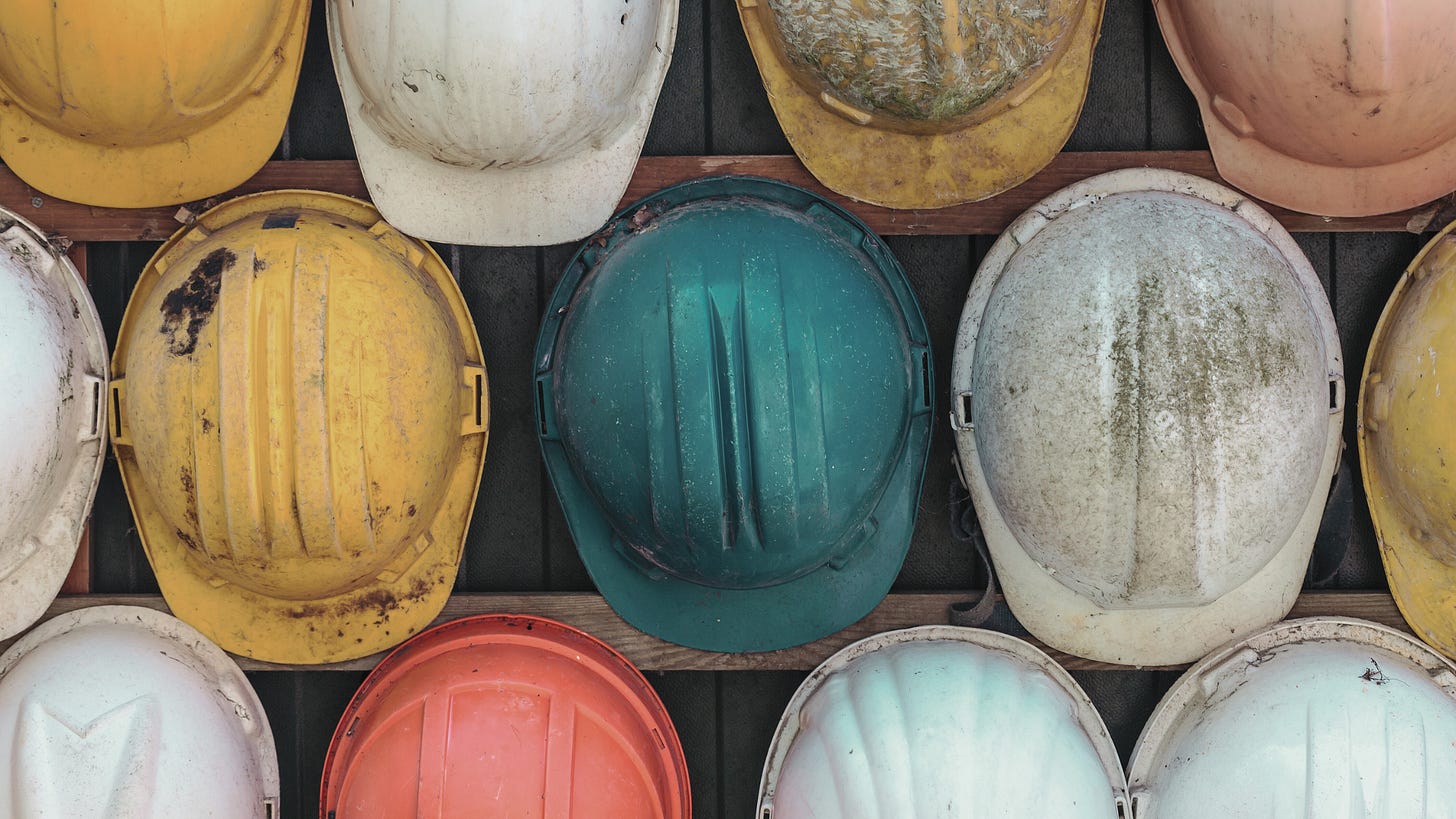 A collection of worn construction hard hats in various colors, including yellow, orange, white, and teal, arranged in a grid pattern on a wooden background. The helmets display scratches, dirt, and signs of use, emphasizing their functionality and history.