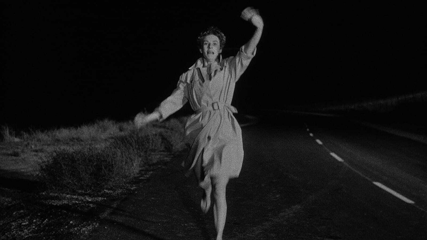 Movie still from Kiss Me Deadly. A woman runs down a road at night, waving her arms in fear