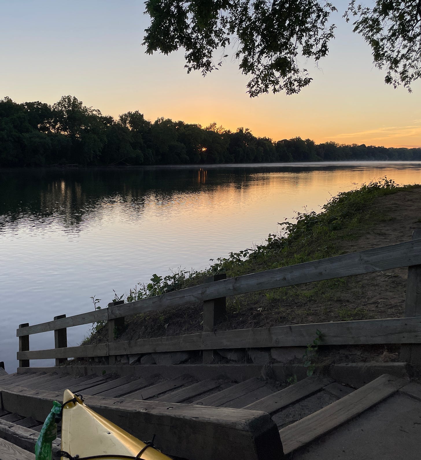 foreground: wooden boat slide down to river; distance - rosy gold sky near the horizon that reflects in the river; sun glowing behind trees on opposite side of river bank.