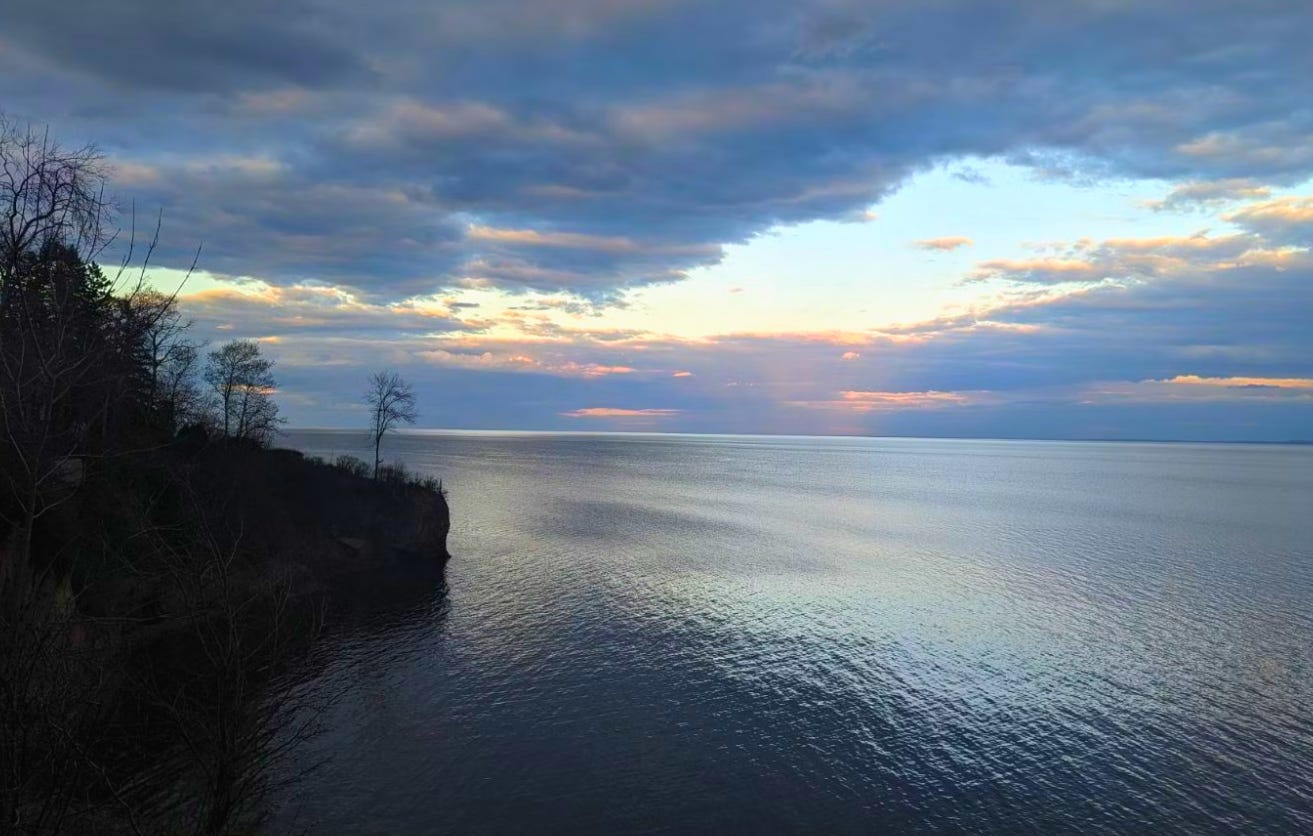 A beautiful photo of Lake Superior. It is dusk and the clouds are tinted pink. The water has just the faint impression of ripples on the surface and there are trees and a rocky cliff on the left side.