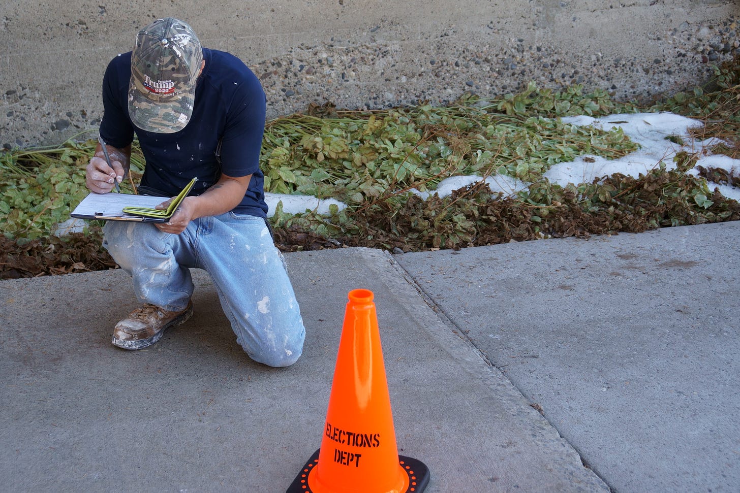 A man in a camouflage Trump hat in jeans, t-shirt and boots dotted with white paint or plaster kneels down while filling out a form on a clipboard.