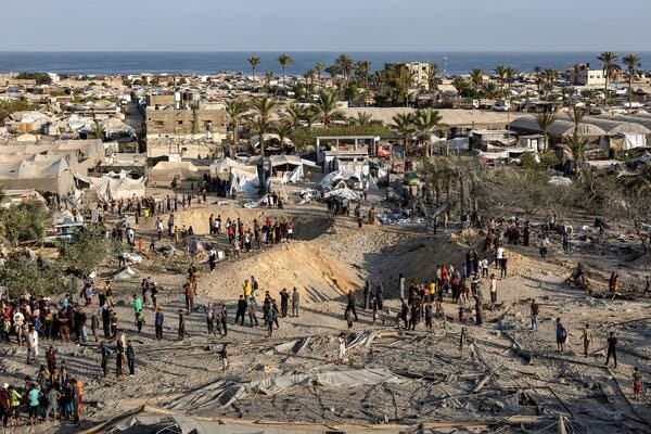 People clustering around two large craters in a sandy area surrounded by tents and low-rise buildings near the seaside. 