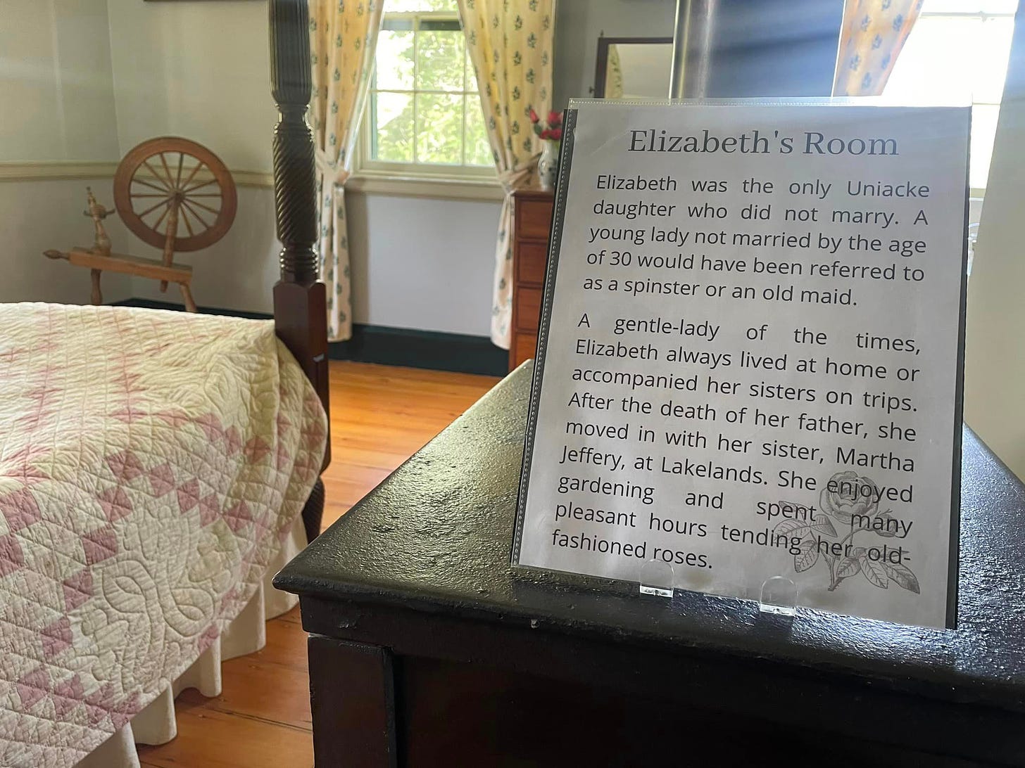 A sign sits on top of a black cabinet in a bedroom next to a four poster bed and a spinning wheel in the corner of the room.