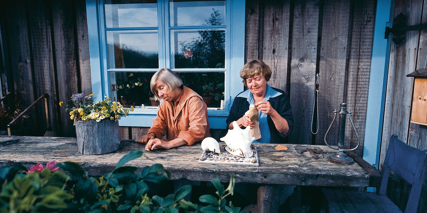 Sitting at a long rustic wood table outside of a wood-plank cottage with sky-blue painted trim, two older white women sit side-by-side doing art.