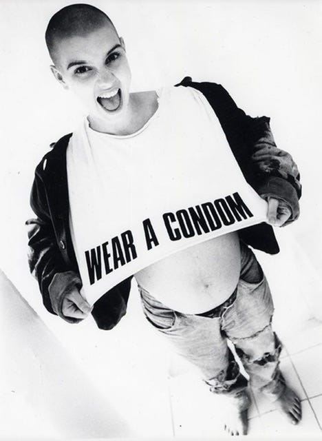 Sinéad O’Connor, foto in bianco e nero. Indossa un giaccone grosso e dei jeans strappati, una maglietta bianca con la scritta “Wear a condom, metti un preservativo” e sotto si intravede il suo pancione (era incinta).