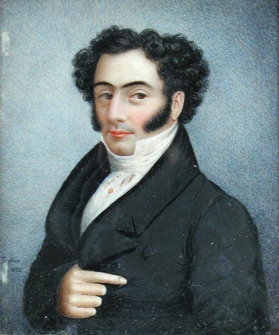 A portrait of a young gentleman in an 1820s-style black suit with a high-collared white shirt. He has black curly hair, sideburns and dark eyebrows. He looks directly at the viewer and holds one hand in front of him on the breast of his jacket.