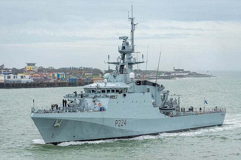 A small, gray British warship cruises through gray waters beneath a cloudy sky on the English coast, with a jetty and boardwalk visible in the background.