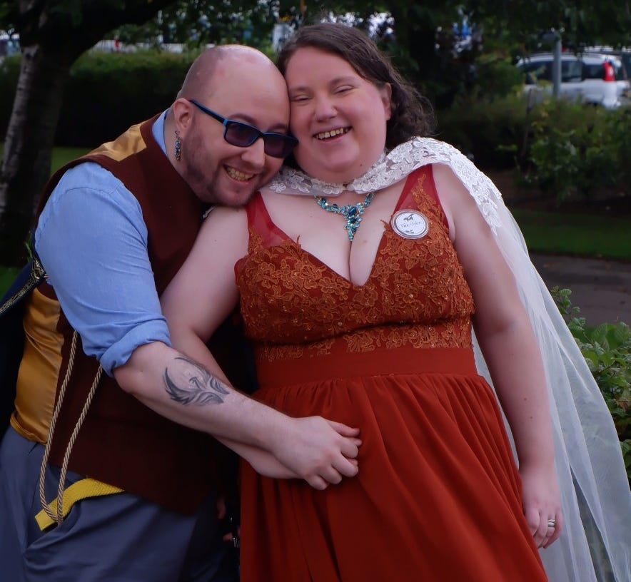 A photo of me (left) and Cuddles (right) outside at our wedding ceremony. I’m bald-headed with a short beard, wearing glasses, a burnt orange waistcoat over a blue shirt, and a grin, resting my chin on Cuddles’ shoulder and holding her right hand. Cuddles wears her brown hair down, a long white train, and a burnt orange dress. She is grinning at the camera, her face resting against mine.