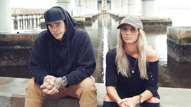 A screenshot of Frankie Holmes (left), wearing a black hoodie and flat-brim baseball cap. He is absolutely covered in tattoos. He sits with his hands clasped in his lap. To his right sits Allie Severino, a young blonde woman, also dressed in all black. They look candidly into the camera as they sit on the side of what appears to be the underpass of a bridge.