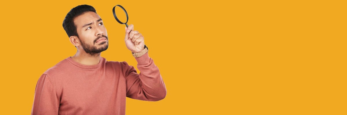 A man in a salmon-colored sweater examines something closely with a magnifying glass against a yellow background.