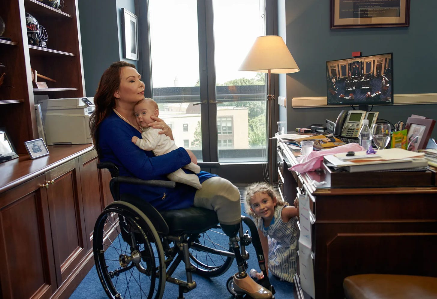 Asian woman with dark hair sits in a wheel chair. She wears a blue dress and holds a baby. She wears an artificial left leg and sits before a brown desk. A child peaks out from under the desk.